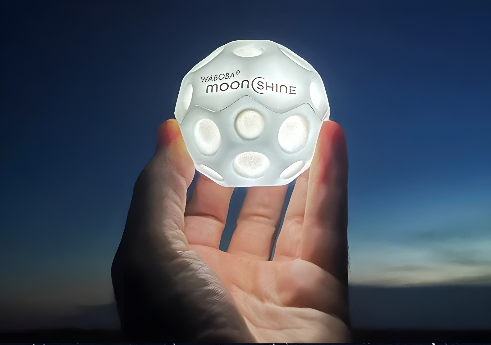 A hand holding a glowing white Waboba Moon Ball against a twilight sky.