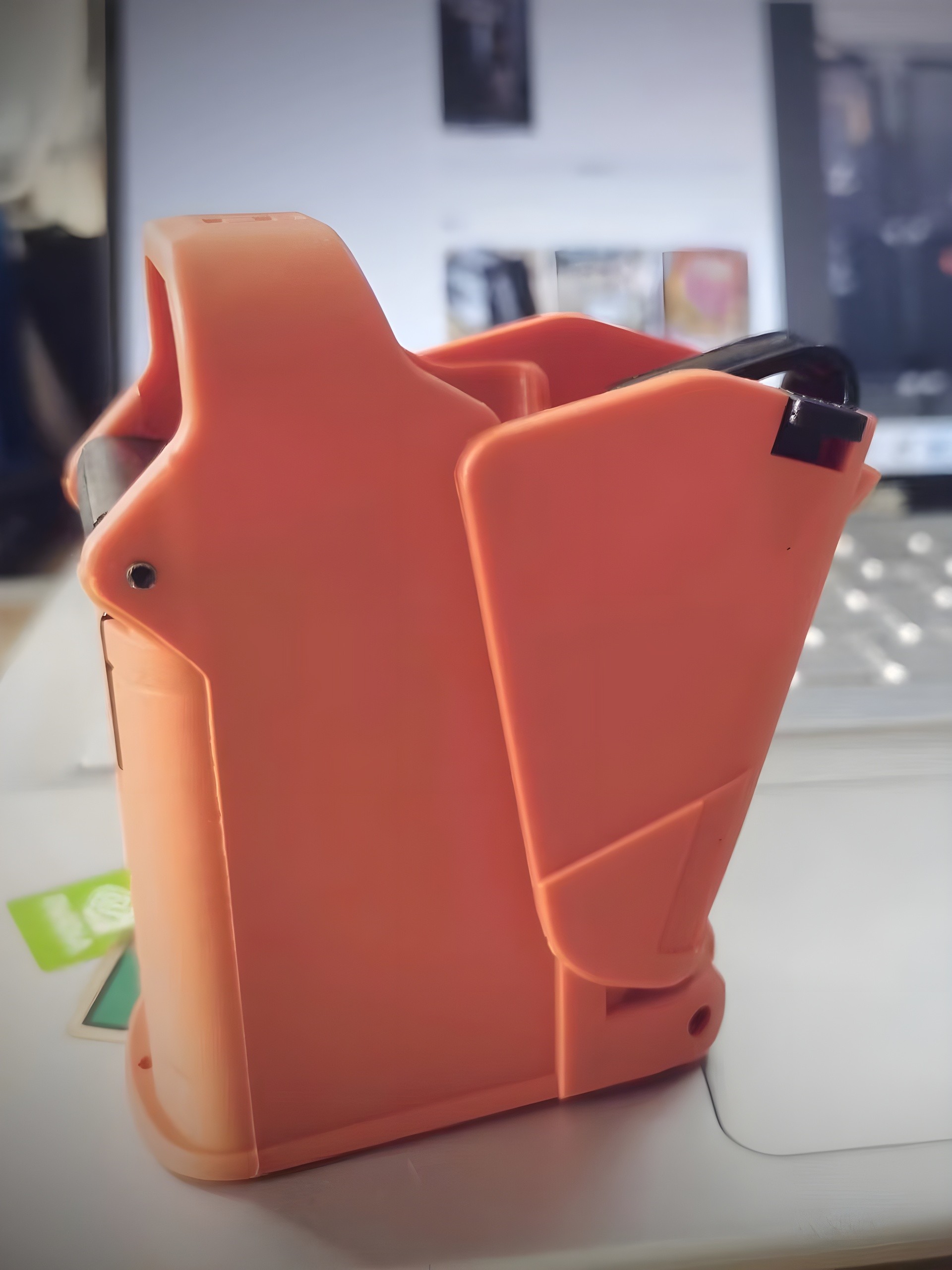 Close-up side view of orange SCETAC pouch on desk, showing asymmetric profile, retention gate, and mounting rivets — background includes laptop keyboard and blurred screen.