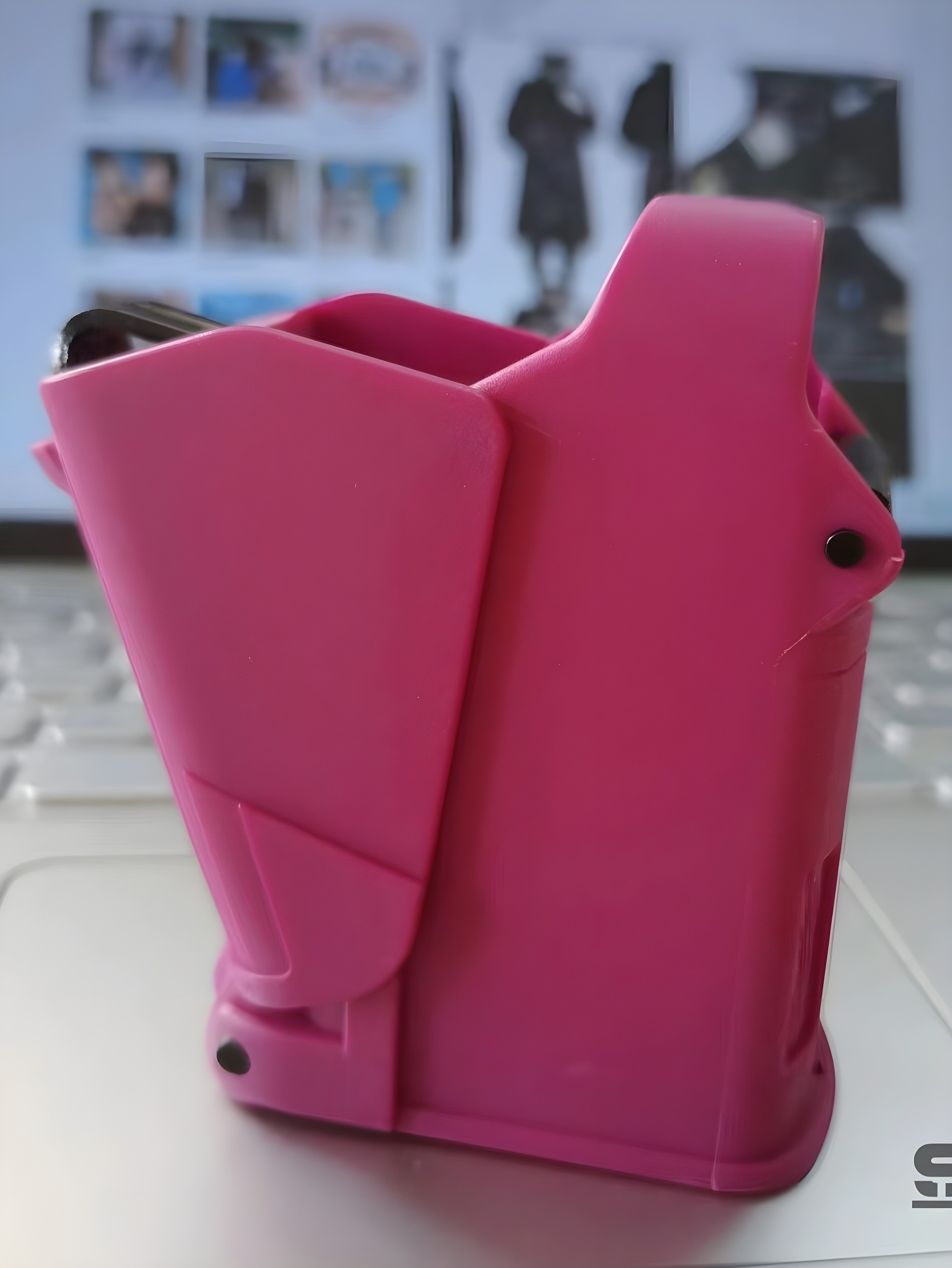 Close-up side view of bright pink SCETAC pouch, highlighting smooth contours, matte finish, and precise rivet placement — placed on laptop surface with blurred photo grid in background.